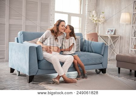 Kissing Mother. Loving Little Girl Feeling Amazing While Kissing Her Pregnant Mother Sitting On Sofa