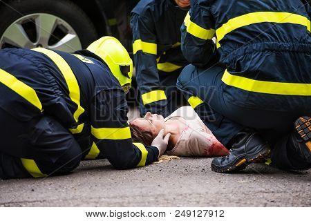 Three Unrecognizable Firefighters Helping A Young Injured Woman Lying On The Road After An Accident.