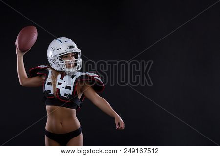 Sportive Serious Woman In Helmet Of Rugby Player Holding Ball In Stuio On Dark Background. Girl Spor