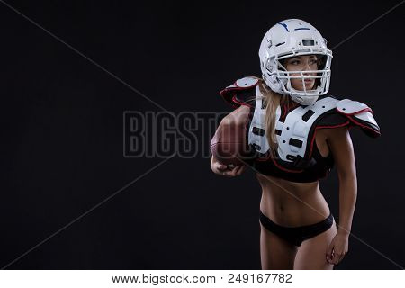 Sportive Serious Woman In Helmet Of Rugby Player Holding Ball In Stuio On Dark Background. Girl Spor