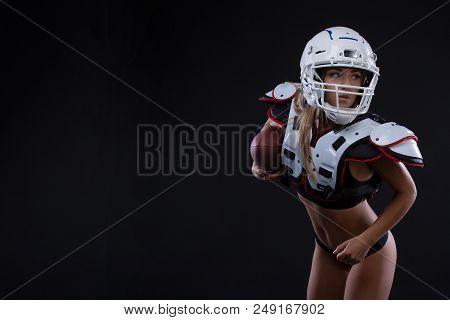 Sportive Serious Woman In Helmet Of Rugby Player Holding Ball In Stuio On Dark Background. Girl Spor