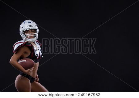 Sportive Serious Woman In Helmet Of Rugby Player Holding Ball In Stuio On Dark Background. Girl Spor