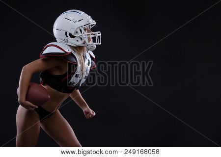 Sportive Serious Woman In Helmet Of Rugby Player Holding Ball In Stuio On Dark Background. Girl Spor