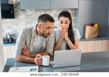 Portrait Of Shocked Married Couple Looking At Laptop Screen Together At Counter In Kitchen