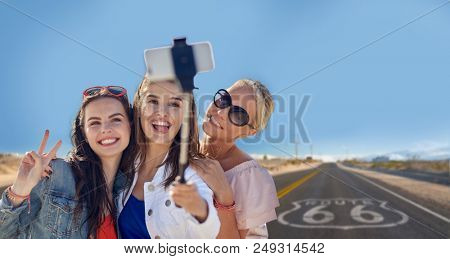summer vacation, holidays, travel, technology and people concept- group of smiling young women taking picture by smartphone on selfie stick over us route 66 background