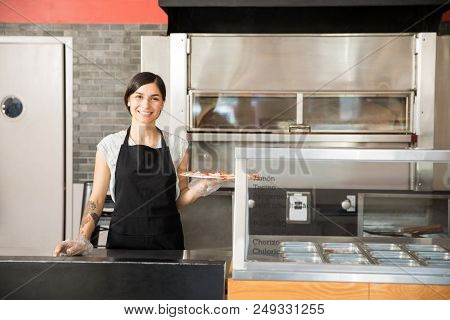 Portrait Of Smiling Chef Wearing Black Apron And Hand Gloves Showing Raw Pepperoni Pizza In Commerci