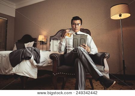 Successful Vietnamese Businessman Drinking Glass Of Whiskey And Reading Information On Tablet Comput