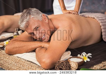Senior man relaxed in spa while having a back massage. Happy mature man feeling relaxed and looking at camera. Male beauty therapy concept.