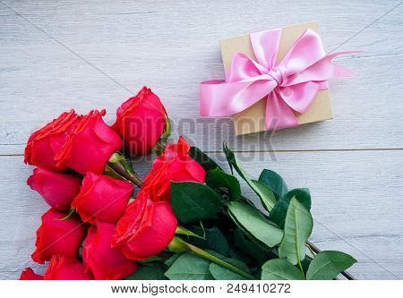 Preparing A Gift, On A Wooden Table, With Roses. Female Hand Holding A Gift, Wrapped With Ribbon, On