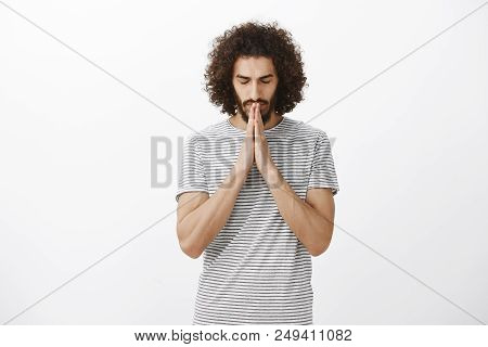 Portrait Of Focused Believing Hispanic Guy With Beard And Afro Hairstyle, Closing Eyes, Holding Hand