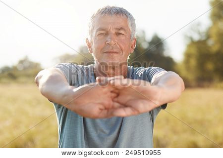 Elederly Grey Haired Wrinkled Male Stretches Hands, Dressed In Casual Outfit, Does Sport Outdoor On 