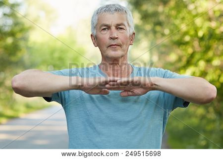 Confident Grey Haired Male Stretches Hands, Does Physical Exercise Or Workout In Open Air, Has Self 