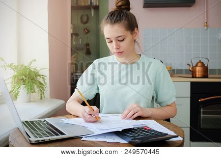 Indoor Picture Of Young Good-looking Female Sitting At Kitchen Table Full Of Papers With Laptop Lyin