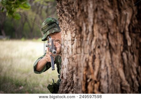 Special Forces Soldier Or Private Millitary Holding Gun Aiming Behind A Tree At Field Area.