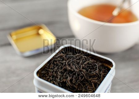 Black Loose Tea In Metal Tin, With Blurred Cup Of Hot Amber Drink In Background.