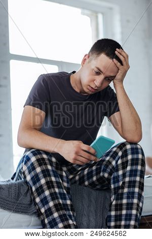 Disillusioned. Miserable Young Man Sitting At Home And Holding His Phone