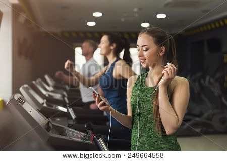 Young Woman With Smartphone Running On Treadmill In Gym. Slim Girl Jogging In Fitness Club, Listen M