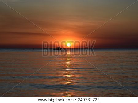 A Cruising Sail Boat Passes Majestically Beneath The Enchanting Sunset. The Sun Casts Orange Shades 