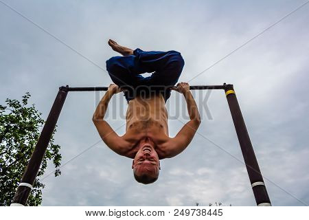 A Man With A Bare Torso In Blue Pants Upside Down Doing His Morning Exercises On A Horizontal Bar Ag