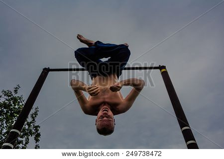 A Man With A Bare Torso In Blue Pants Upside Down Doing His Morning Exercises On A Horizontal Bar Ag