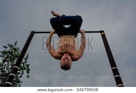 A Man With A Bare Torso In Blue Pants Upside Down Doing His Morning Exercises On A Horizontal Bar Ag