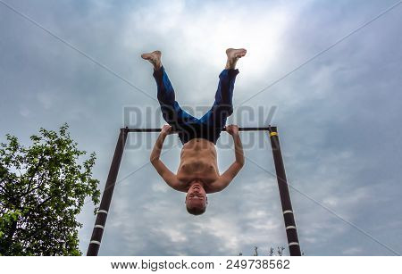 A Man With A Bare Torso In Blue Pants Upside Down Doing His Morning Exercises On A Horizontal Bar Ag