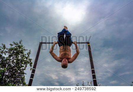 A Man With A Bare Torso In Blue Pants Upside Down Doing His Morning Exercises On A Horizontal Bar Ag