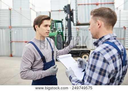 Misunderstanding young cargo employee in workwear gesturing while asking foreman about loading and explaining characteristics of forklift