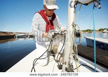 erious concentrated stylish handsome mature sailor in striped sweater and cap checking anchorage of ropes and standing on one knee on yacht deck