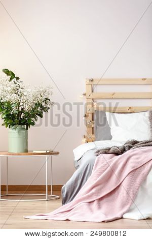Flowers On Table Next To Bed With Pink Blanket And Wooden Headboard In Bedroom Interior. Real Photo