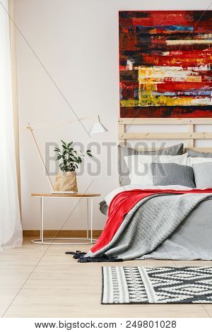 Grey And Red Blanket On Bed Next To Table With Lamp And Plant In Bedroom Interior With Painting. Rea