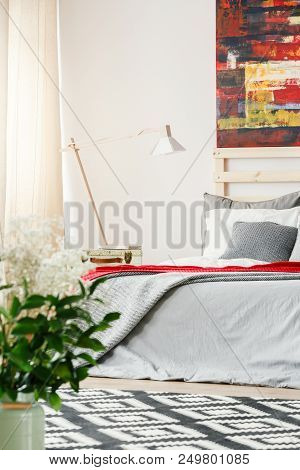 Lamp Next To Grey Bed In Bright Bedroom Interior With Red Painting And Flowers. Real Photo
