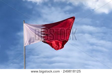 Real Flag Of The Country Qatar Waving In The Wind Against The Background Of A Blue Sky And Clouds.