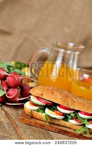 Large Baguette Sandwich Cut In Half Filled With Egg, Arugula Salad, Tomatoes And Radish In Backgroun