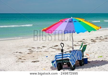 Colorful Beach Umbrella With A Wagon, Striped Towel, Chair And Toys On The Beach