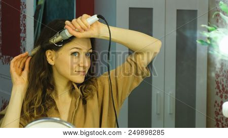 Young Beautiful Girl Looks In The Mirror And Curls Her Hair On A Curling Iron In The Dressing Room, 