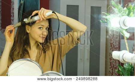 Young Beautiful Girl Looks In The Mirror And Curls Curls On The Curling Iron In The Dressing Room.