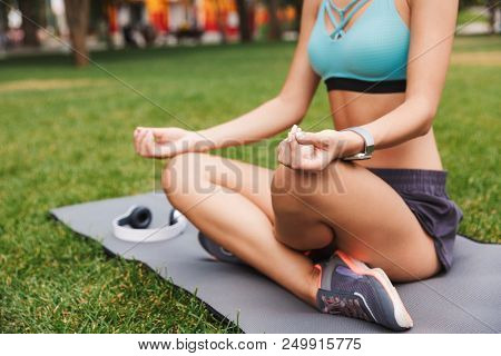 Cropped image of relaxed young sportswoman dressed in sportswear meditating while sitting in a yoga position on a fitness mat on grass outdoors