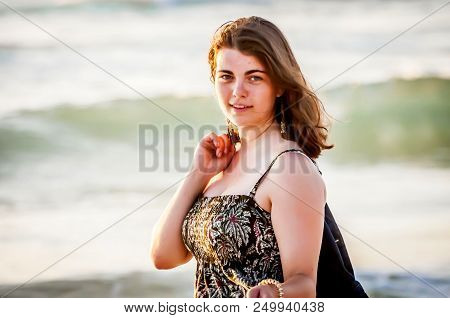 Nice Looking Cute Caucasian Girl In A Long Gaudy Dress At The Seaside, With Sea Waves On The Backgro