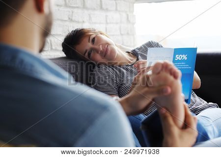 Young Hispanic Couple Laying On Couch At Home, Holding A Travel Flyer And Planning Next Trip. The Bo
