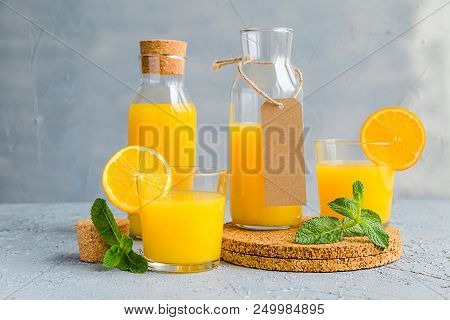 Orange Freshly-squeezed Juice In A Glass And Bottles On Grey Table.