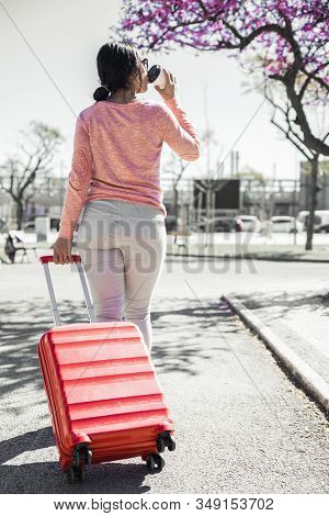 Back View Of Young Woman Pulling Trolley Case Outdoors. Lady Drinking Coffee And Walking With City V