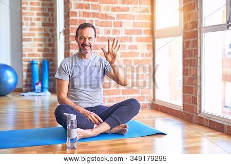 Middle age handsome sportman sitting on mat doing stretching yoga exercise at gym showing and pointing up with fingers number five while smiling confident and happy.