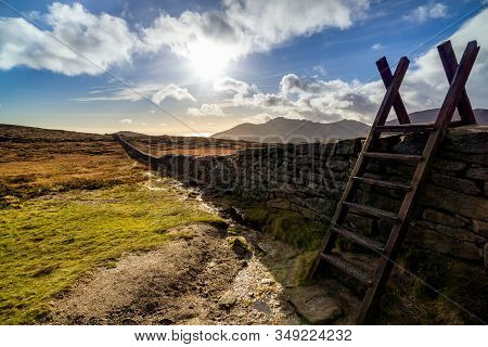 Mourn Wall With Ladder On The Bank Of Slieve Donard Mountain With Blue Sky, White Clouds And Sunrays