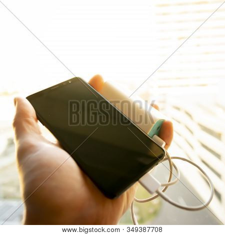 Hand Holding Black Smartphone (mobile Pohone) Which Is Being Charged With A Battery Power Bank Again