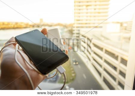 Hand Holding Black Smartphone (mobile Pohone) Which Is Being Charged With A Battery Power Bank Again