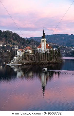 Island Church At Lake Blend In Slovenia During Sunrise. A Pletna Boat Is Transporting Tourists.