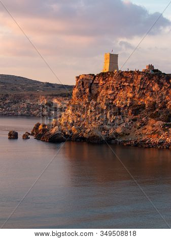A Tower Stands On The Cliff Edge By The Sea. The Cliff Glow A Warm Orange During Sunset