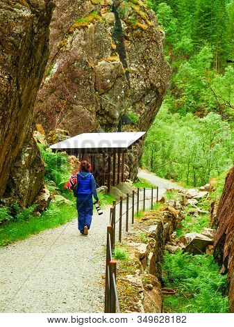 Tourist Woman With Norwegian Flag And Camera Walking Trough Mountains Path In Allmannajuvet Area Sau