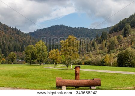 Landscape With Wooden Drinking Fountain, Green Meadow, Multi Colored Trees, Mountain Range With Fore